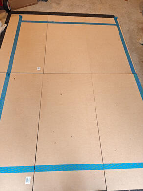 MDF laid out for measuring six MDF panels, laid out on the floor. Blue masking tape has been used to measure the dimensions of the wooden flats, for cutting.